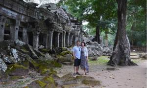 A couple, standing in front of the Ta Prohm Temple in Cambodia, which is overgrown by trees.
