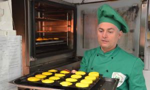 A chef, wearing a green hat and uniform, holds a tray of bright yellow souffles, preparing to put them in an oven.