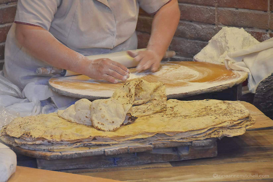 A woman rolls out a ball of lavash flatbread dough on top of a circular surface.
