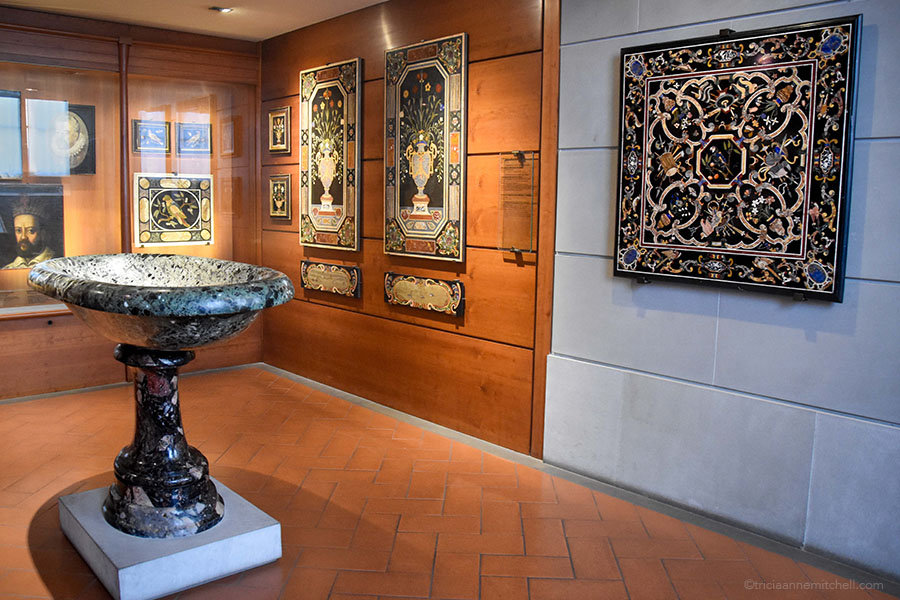 A large mostly back stone fountain sits in the middle of a museum display alongside wallpaintings created via the Florentine marquetry (Florentine mosaic) technique.