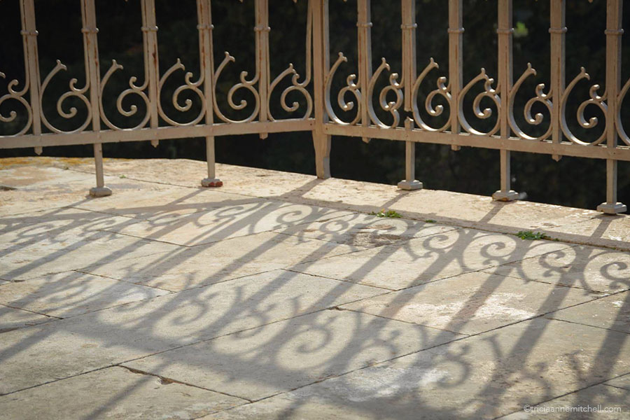 The swirly embellishments of an iron railing cast shadows on a stone walkway